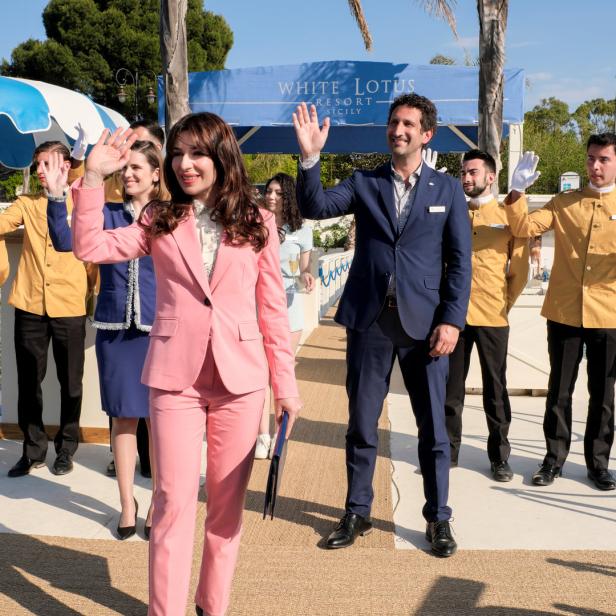 Eine Gruppe von Menschen, darunter eine Frau in einem pinkfarbenen Anzug, winkt vor dem „White Lotus Resort Sicily“.