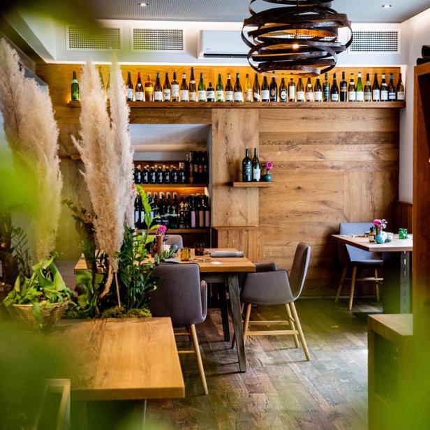 Innenansicht eines Restaurants mit Holztischen, Stühlen und einer Wand mit vielen Weinflaschen.