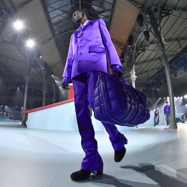 Ein Model in einem lila Anzug und mit einer passenden Tasche auf einem Laufsteg.
