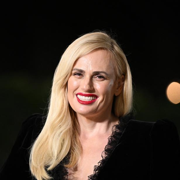 Rebel Wilson lächelt in einem schwarzen Kleid mit roten Lippen.