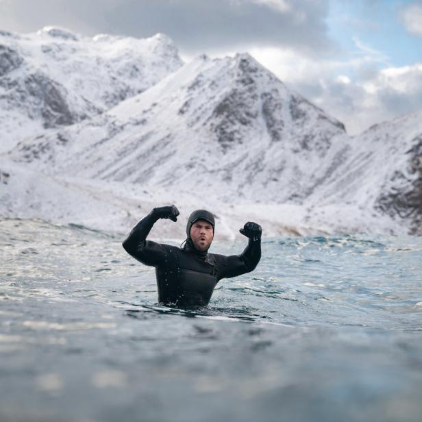 Ein Mann im Neoprenanzug steht im Wasser vor einer schneebedeckten Berglandschaft und ballt die Fäuste.