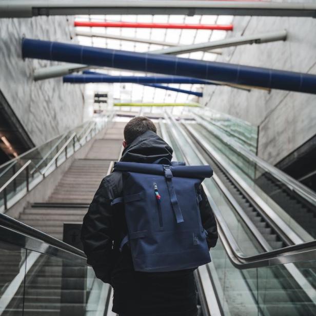 Ein Mann mit blauem Rucksack fährt eine Rolltreppe in einem modernen Gebäude hinauf.
