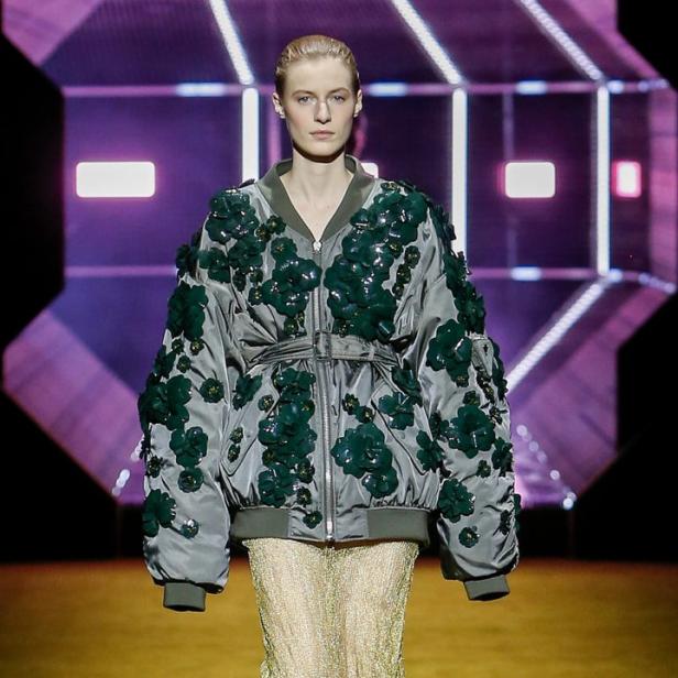 Ein Model auf dem Laufsteg trägt eine graue Bomberjacke mit grünen Blumenapplikationen und einen goldenen Rock.