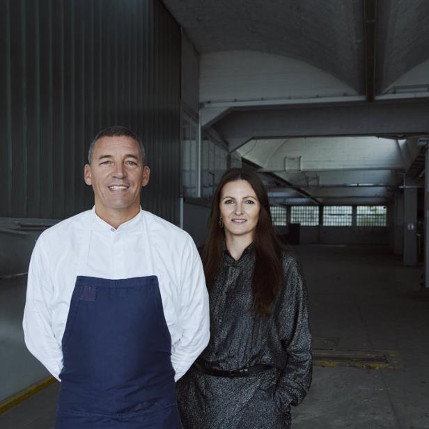 Ein Koch mit Schürze und eine Frau stehen in einem industriell anmutenden Raum.