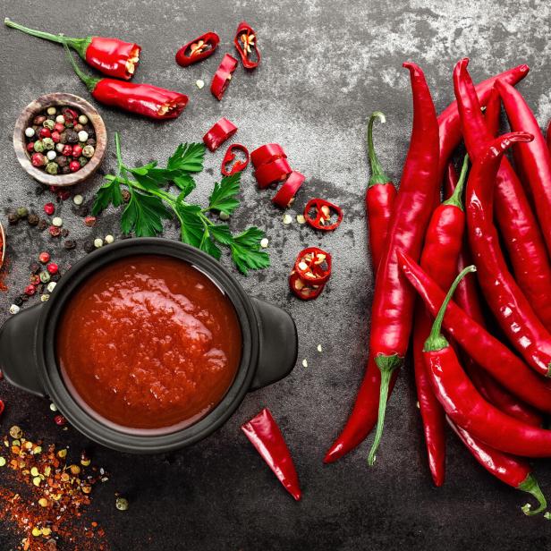 Rote Chilischoten, Gewürze und eine Schale mit roter Sauce auf dunklem Untergrund.