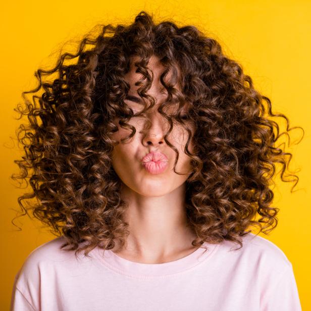 Eine Frau mit lockigem Haar und rosa Lippen vor einem gelben Hintergrund.
