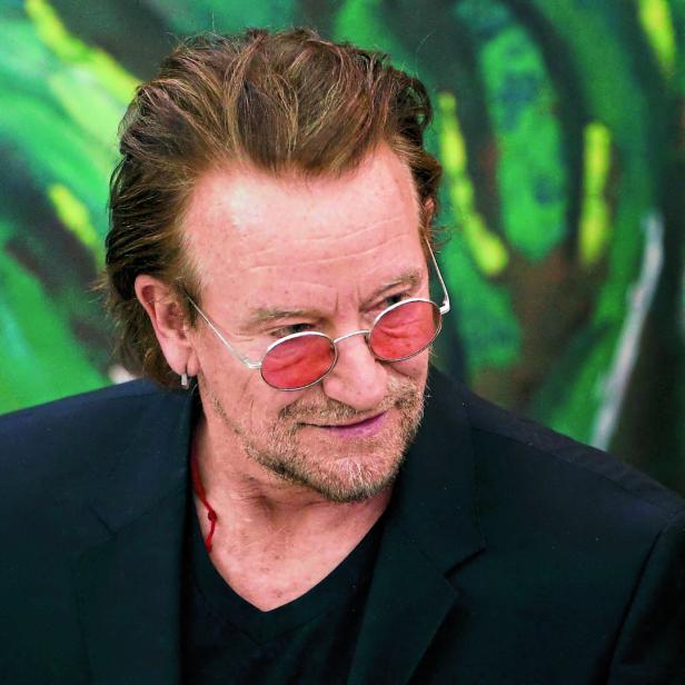 Bono von U2 mit getönter Brille vor einem farbenfrohen Hintergrund.