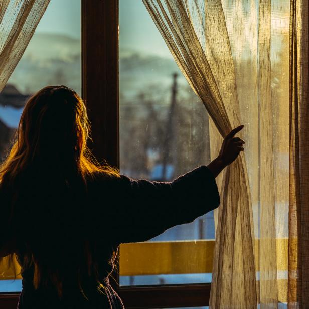 Eine Frau im Morgenmantel öffnet die Vorhänge und blickt aus dem Fenster.