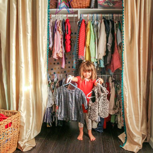 Ein kleines Mädchen steht vor einem offenen Kleiderschrank und hält zwei Kleidungsstücke hoch.