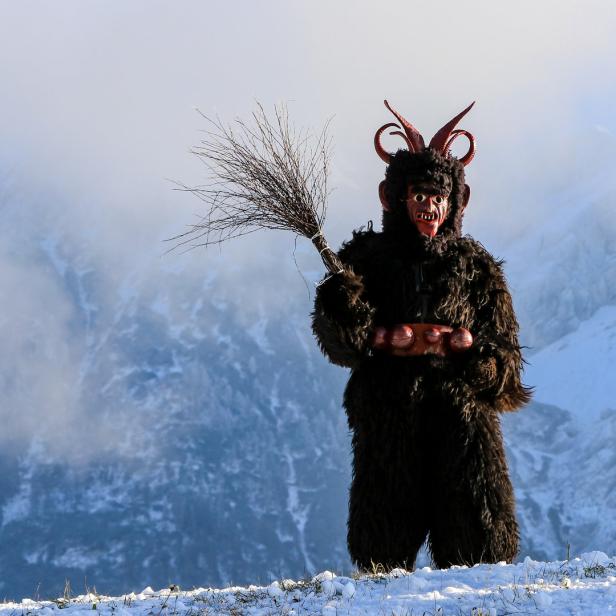 Ein Krampus mit Fellkostüm, Maske und Besen steht vor einer verschneiten Berglandschaft.