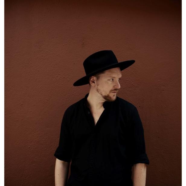 Ein Mann mit Hut und schwarzem Hemd vor einer braunen Wand.