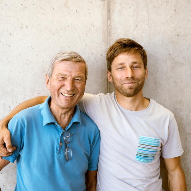 Ein älterer Mann und ein jüngerer Mann stehen lächelnd nebeneinander vor einer Betonwand.