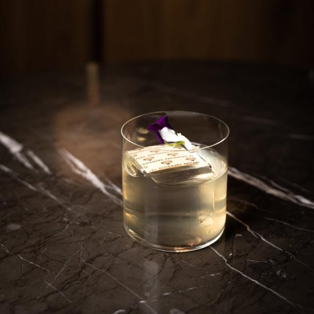 Ein Cocktail mit einem großen Eiswürfel und einer lila Blüte auf einem Marmortisch.