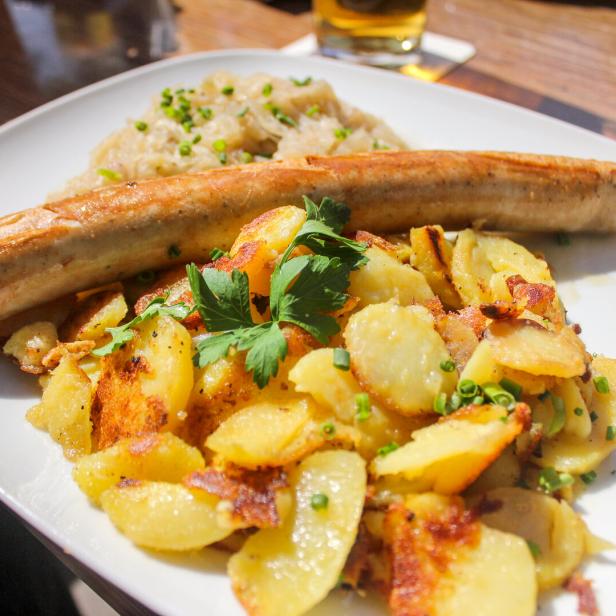 Eine Bratwurst liegt auf Bratkartoffeln und Sauerkraut auf einem Teller.