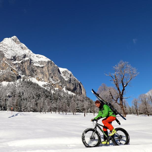Ein Mann fährt mit einem Fatbike und Skiern auf dem Rücken durch eine verschneite Landschaft.