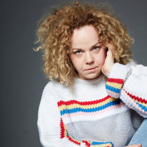 Eine Frau mit lockigem Haar sitzt in Jeans und einem bunten Pullover.