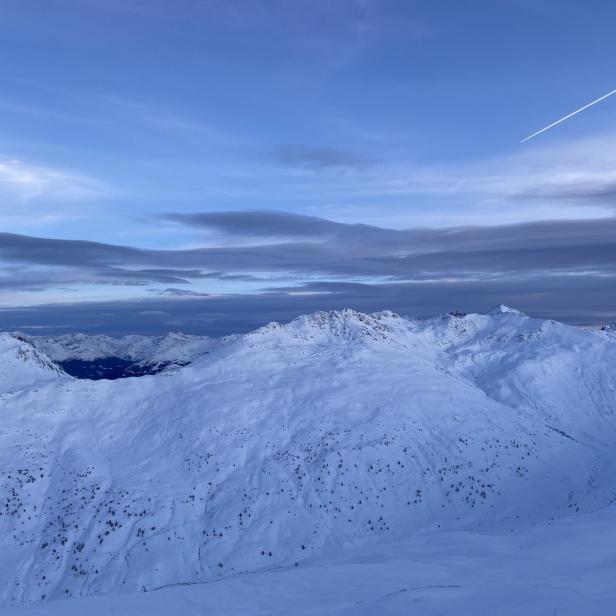 Eine verschneite Berglandschaft unter einem Himmel mit Wolken, einem Flugzeugstreifen und dem Mond.