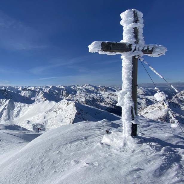 Ein Gipfelkreuz steht auf einem schneebedeckten Berg, im Hintergrund eine Berglandschaft.