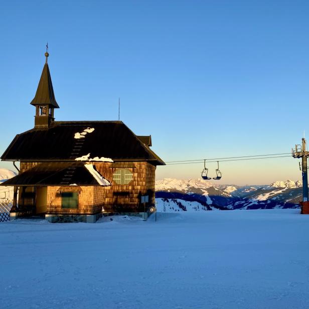 Eine kleine Kapelle steht auf einem schneebedeckten Berg, im Hintergrund eine Liftanlage und weitere Berge.