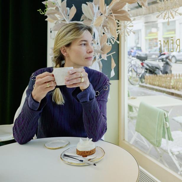 Eine Frau sitzt in einem Café, trinkt Kaffee und blickt aus dem Fenster.