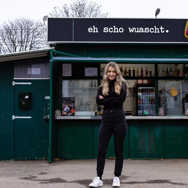 Eine junge Frau steht vor einem Imbissstand mit der Aufschrift „eh scho wuascht“.