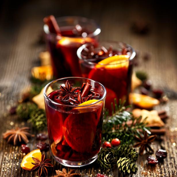 Drei Gläser Glühwein, garniert mit Sternanis, Zimtstangen und Orangenscheiben, auf einem Holztisch.