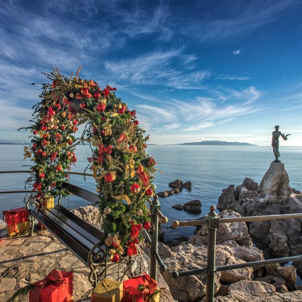 Ein mit Weihnachtsschmuck dekorierter Bogen und Geschenke vor einer Küstenlandschaft mit einer Statue.