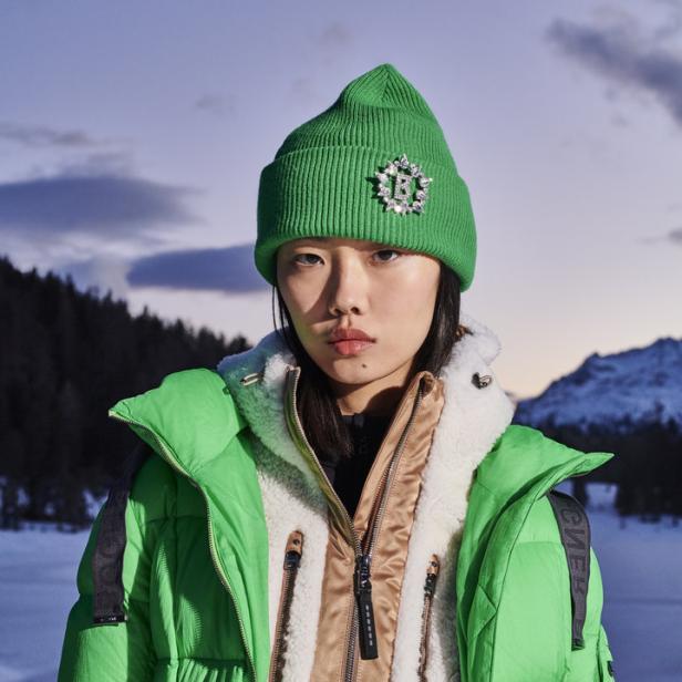 Eine Frau mit grüner Mütze und Jacke vor einer verschneiten Berglandschaft.