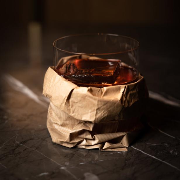 Ein Cocktail mit großem Eiswürfel in einem Glas, das in Papier gewickelt ist.