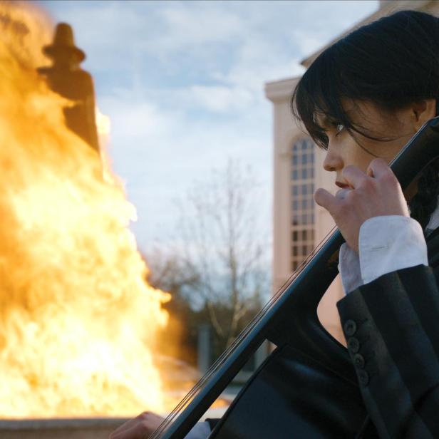 Wednesday Addams spielt Cello vor einem lodernden Feuer.