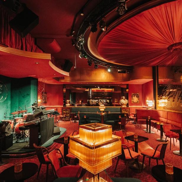 Innenansicht eines eleganten, rot beleuchteten Jazzclubs mit Bühne und Bar.