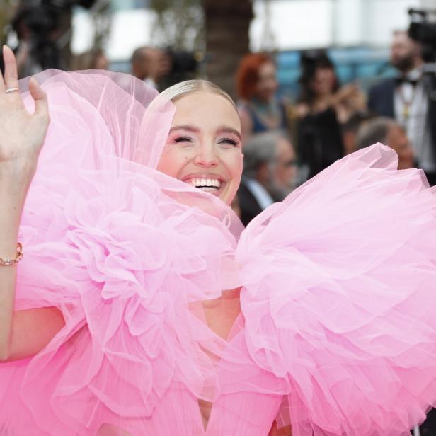 Eine Frau in einem auffälligen, pinkfarbenen Tüllkleid winkt lächelnd.