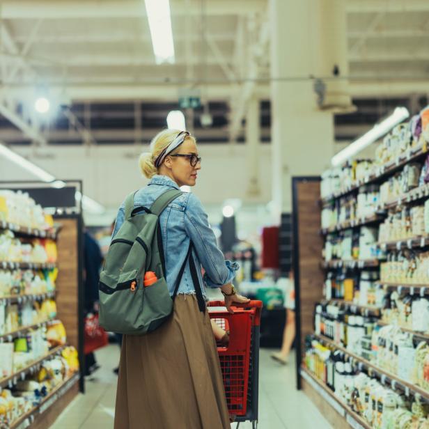 Eine Frau mit Rucksack schiebt einen Einkaufswagen durch einen Supermarkt.