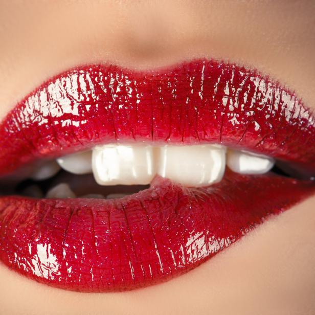 Nahaufnahme von einem Mund mit glänzendem, rotem Lippenstift, der leicht auf die Unterlippe beißt.