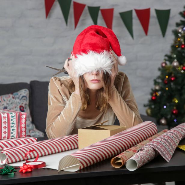 Eine Frau mit Weihnachtsmütze sitzt frustriert vor Geschenkpapier und Schere.