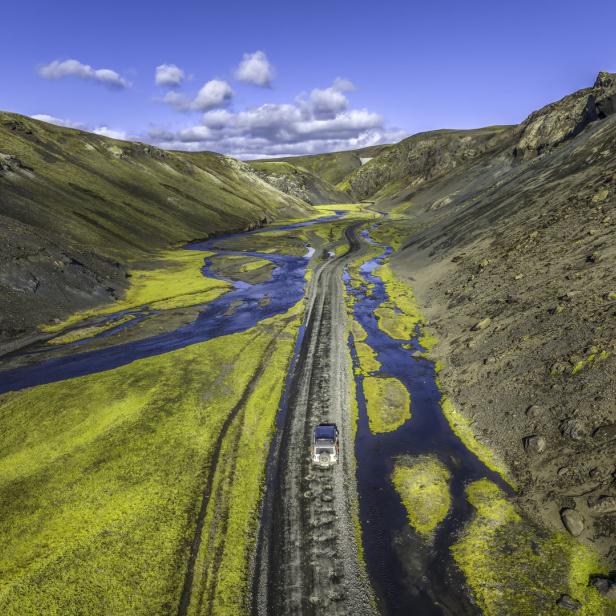 Ein Auto fährt auf einer Schotterstraße durch eine isländische Flusslandschaft.