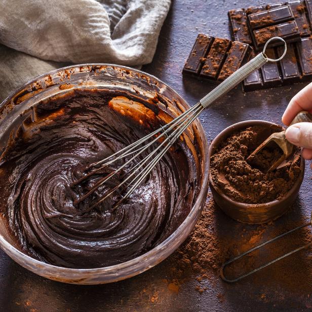 Eine Schüssel mit Schokoladencreme, Kakaopulver, Schokolade, Haselnüsse und eine Hand mit einem Messlöffel.