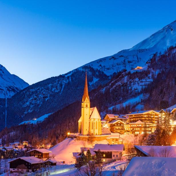 Ein verschneites Dorf in den Alpen mit einer beleuchteten Kirche im Abendlicht.
