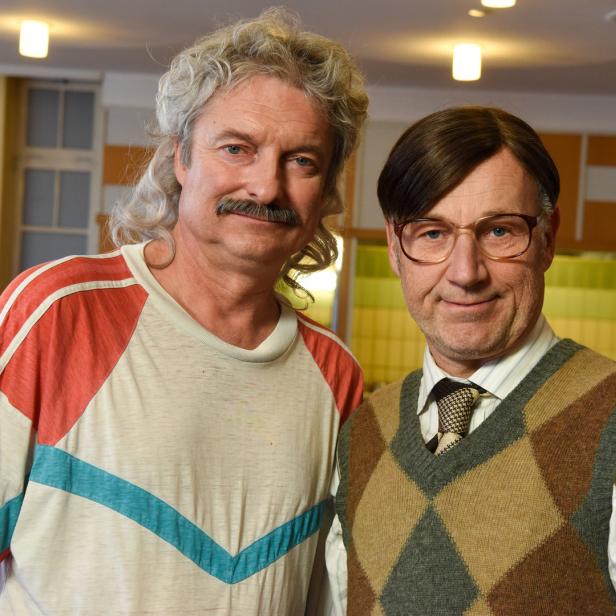 Zwei Männer posieren nebeneinander, der eine mit Schnurrbart und langem, gewelltem Haar, der andere mit Brille und Pullunder.
