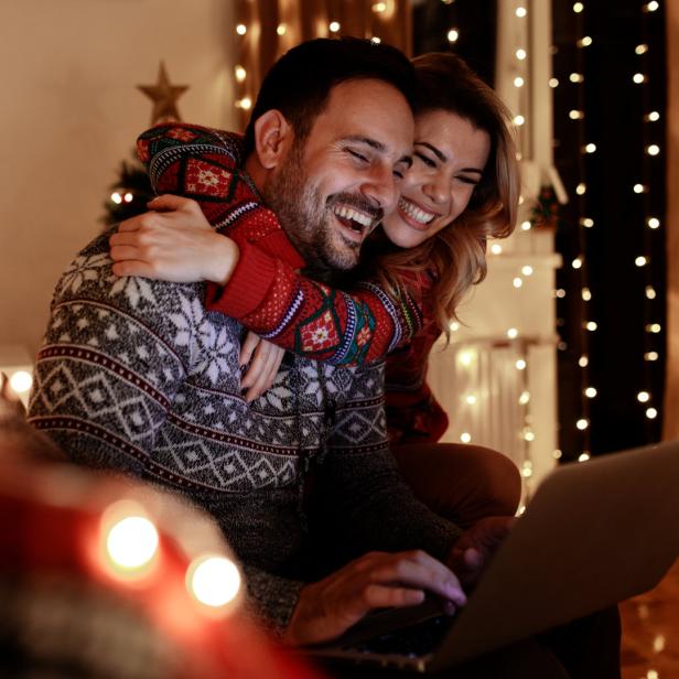 Ein lächelndes Paar mit Weihnachtspullovern sitzt vor einem Laptop, umgeben von Lichtern.