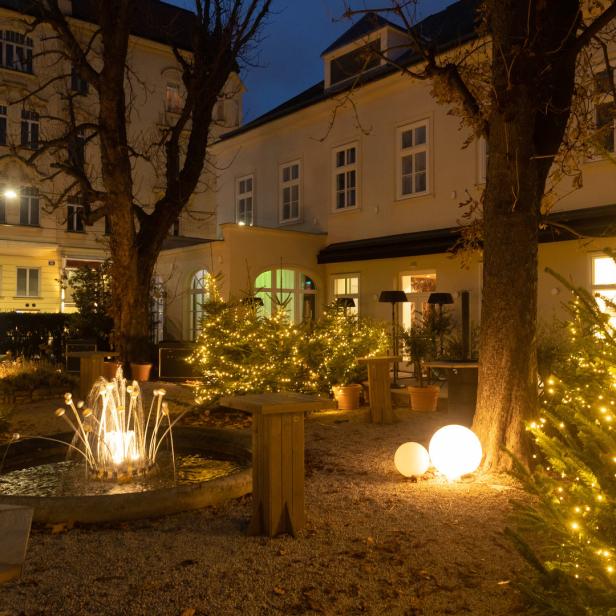 Ein Garten mit beleuchteten Weihnachtsbäumen, einem Brunnen und dekorativen Kugellampen bei Nacht.