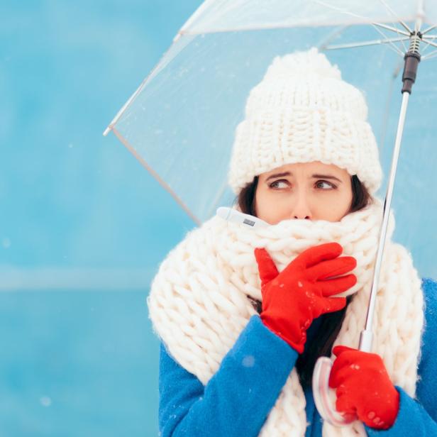 Eine kranke Frau mit Thermometer im Mund hält einen Regenschirm im Schnee.