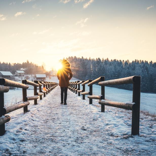 Eine Person geht auf einer schneebedeckten Brücke in Richtung Sonnenaufgang.