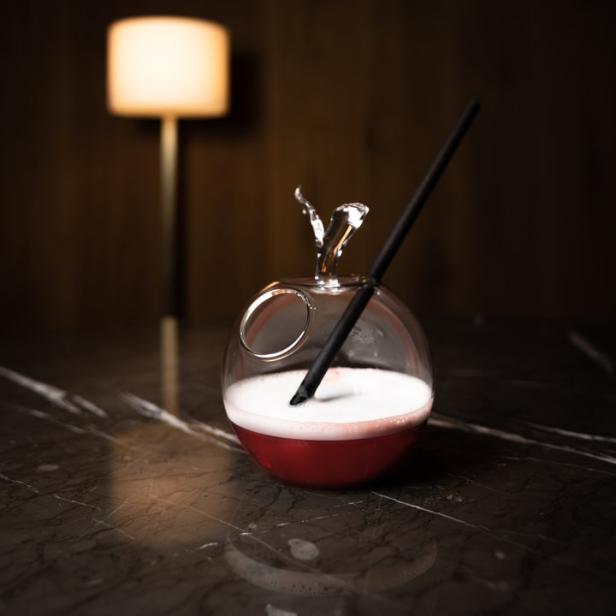 Ein rosafarbener Cocktail in einem Apfel-förmigen Glas mit einem schwarzen Strohhalm auf einem Tisch.