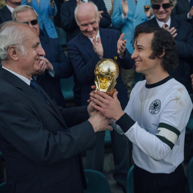 Ein Mann im Anzug überreicht einem Fußballspieler den Weltpokal.