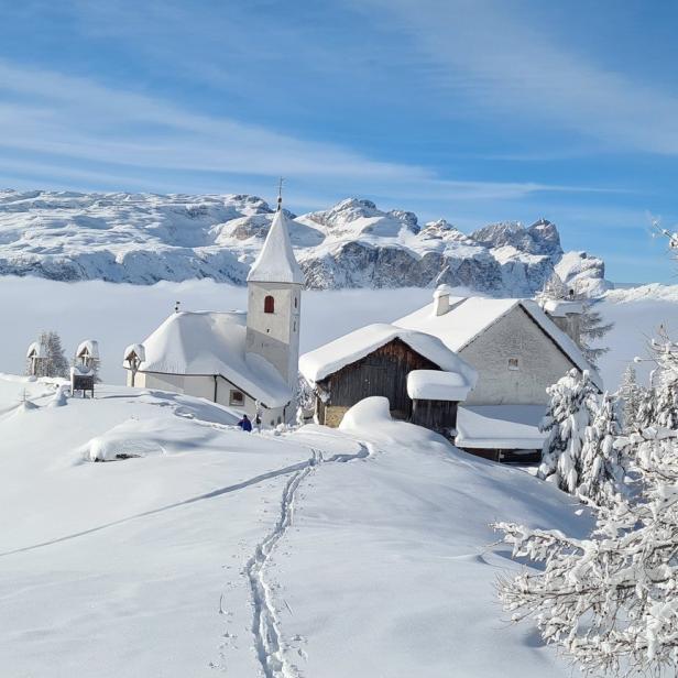 Eine verschneite Kirche und ein Haus auf einem Hügel mit Bergen im Hintergrund.