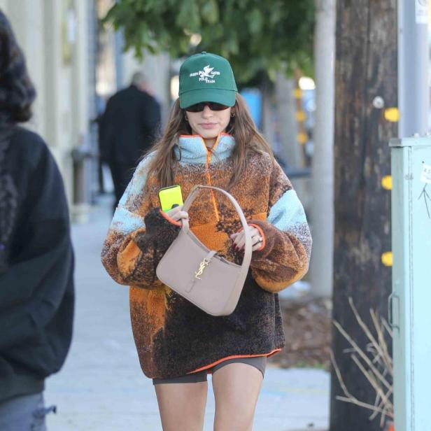 Hailey Bieber mit einer grünen Kappe, einer YSL-Tasche und einem bunten Fleece-Pullover auf der Straße.