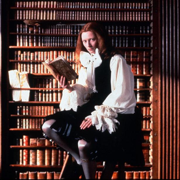 Tilda Swinton sitzt in einer Bibliothek und liest ein Buch.