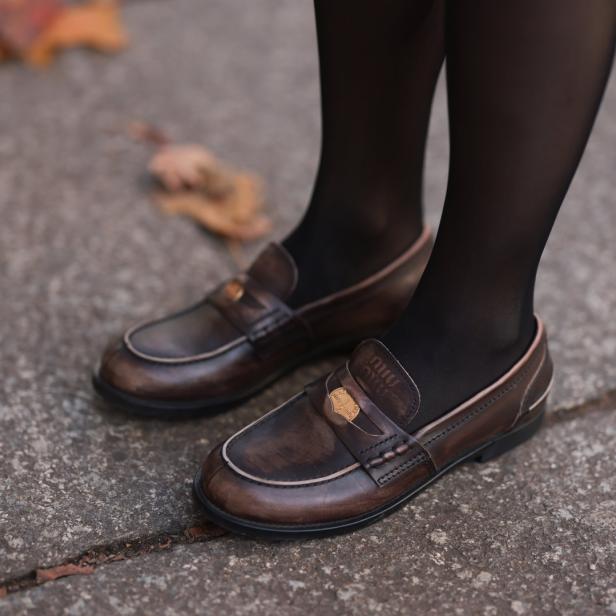 Nahaufnahme von braunen Miu Miu Loafern mit schwarzen Strumpfhosen auf einem Gehweg.