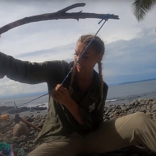Eine Frau sitzt am Strand und spannt einen selbstgebauten Bogen.
